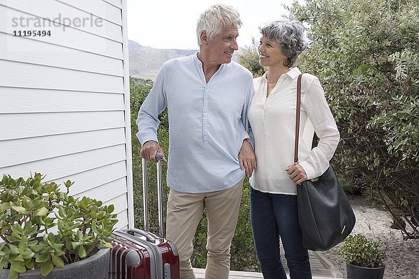 Glückliches Seniorenpaar steht mit Koffer vor dem Haus