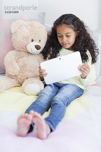 Kleines Mädchen mit einem digitalen Tablett mit Teddybär auf dem Bett