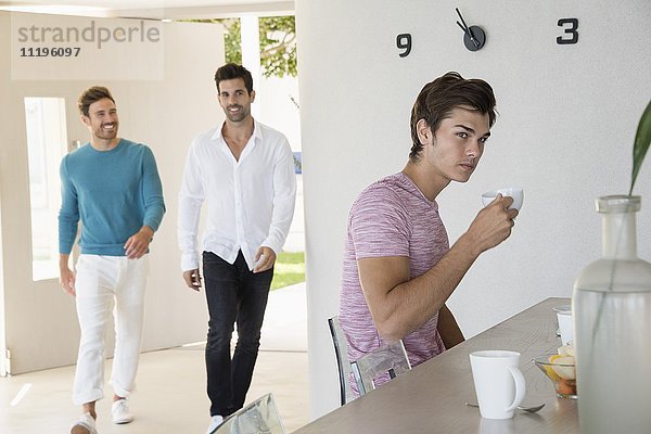 Junger Mann beim Kaffee zu Hause mit seinen Freunden aus dem Hintergrund