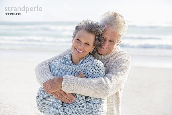 Ein älterer Mann umarmt seine Frau von hinten am Strand.