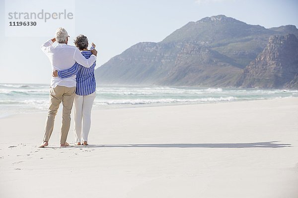 Rückansicht eines Paares  das mit einem Arm um den anderen am Strand steht.