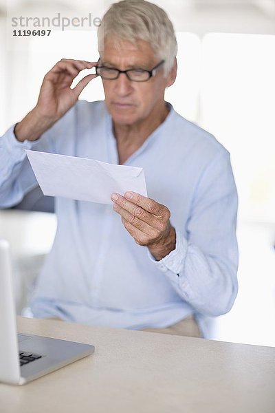 Senior Mann beim Lesen eines Briefes mit Laptop auf dem Tisch