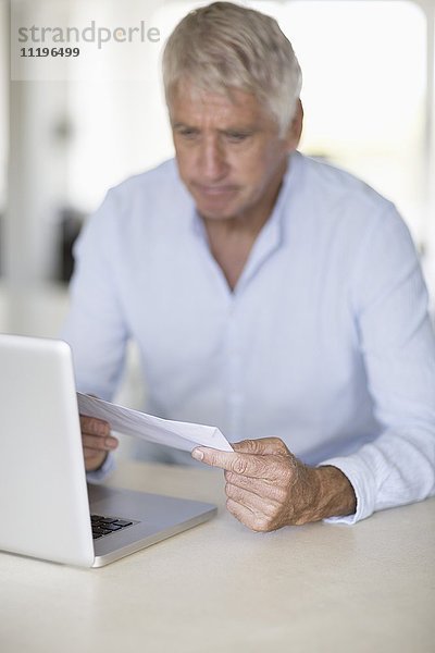 Senior Mann beim Lesen eines Briefes mit Laptop auf dem Tisch