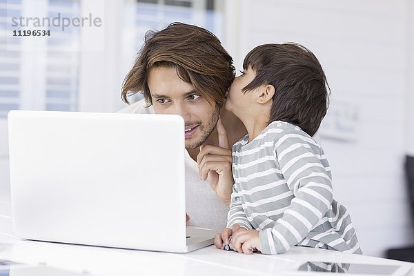 Sohn flüstert Vater ins Ohr  während er den Laptop zu Hause benutzt.