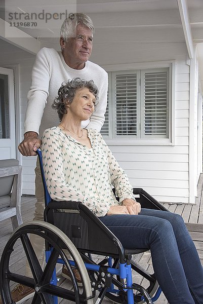 Senior Mann  der seiner Frau im Rollstuhl auf der Veranda assistiert.