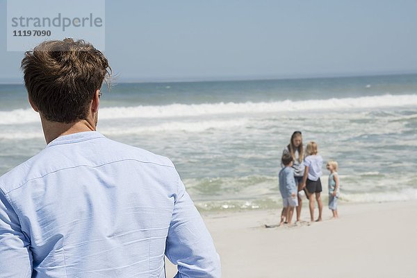 Rückansicht des Mannes mit Blick auf seine Familie am Strand