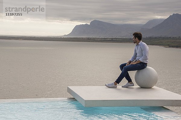 Mann auf Steinkugel am Seeufer sitzend