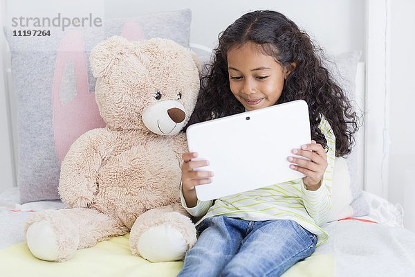 Glückliches kleines Mädchen mit einem digitalen Tablett mit Teddybär auf dem Bett