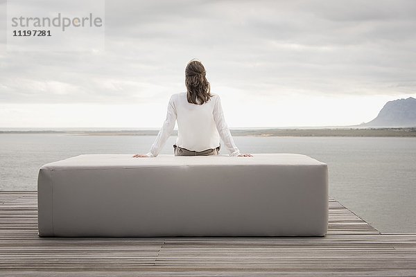 Rückansicht einer Frau  die auf einem Hocker am Seeufer sitzt.