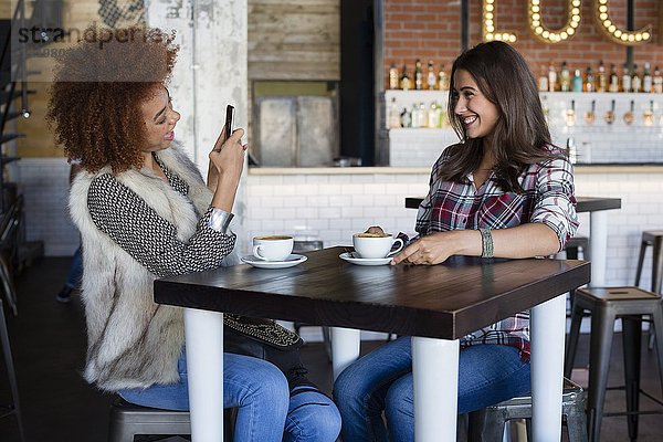 Junge Frau beim Fotografieren ihrer Freundin mit Smartphone im Café