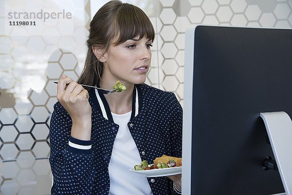 Schöne Frau sitzt vor dem Computer und isst zu Mittag.