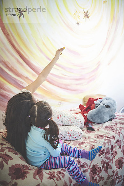 Hispanische Mutter und Tochter auf dem Bett mit Blick auf eine Wandmalerei an der Wand