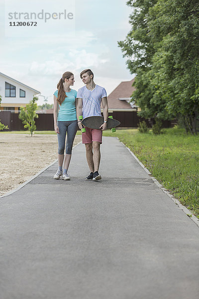 Mann mit Skateboard  der mit seiner Freundin auf einem Spazierweg an Bäumen im Park spazieren geht