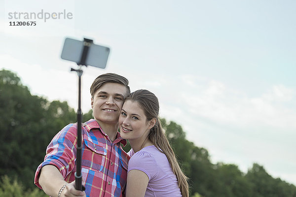 Fröhliches junges Paar nimmt Selfie durch Einbeinstativ im Park gegen den Himmel
