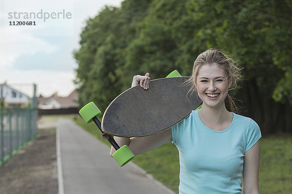 Porträt einer fröhlichen Frau mit einem Skateboard auf einem Fußweg im Park.