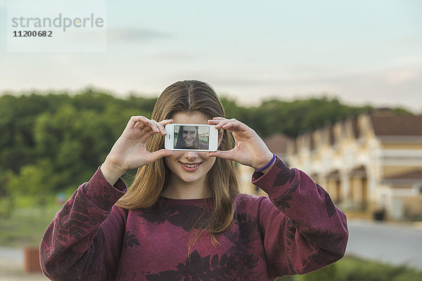 Fröhliche junge Frau zeigt Display von Smartphone mit ihrem Selfie im Freien