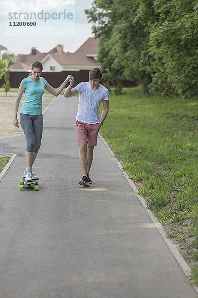 Mann assistiert Freundin beim Skateboardfahren auf dem Fußweg im Park