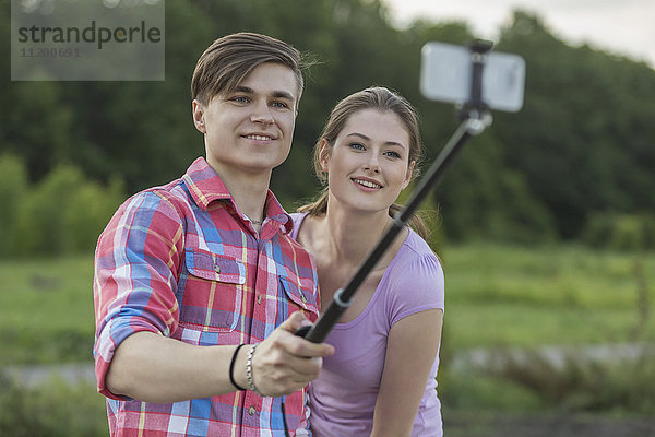 Lächelndes junges Paar nimmt Selfie durch Einbeinstativ am Park gegen Bäume