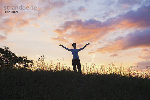 Frau steht mit ausgestreckten Armen auf grasbewachsenem Feld gegen den Himmel bei Sonnenuntergang.