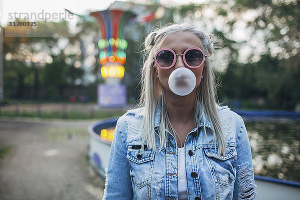 Portrait einer jungen Frau mit Sonnenbrille beim Blasen von Kaugummi im Vergnügungspark