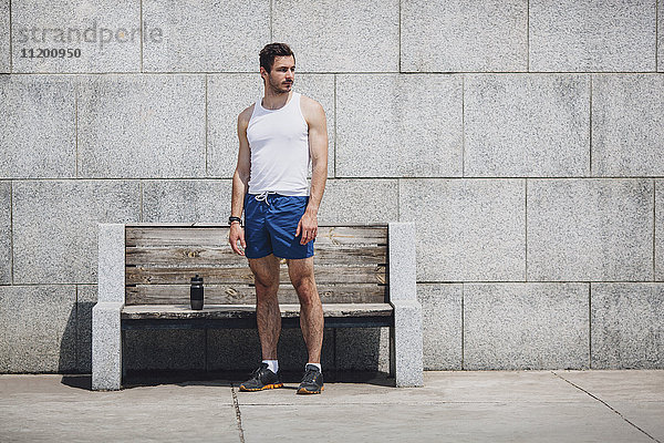 Seriöser Sportler steht an einer Holzbank auf dem Bürgersteig gegen die Wand.