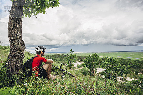 Müde Mountainbiker ruhen unter einem Baum auf einem Hügel gegen den bewölkten Himmel.