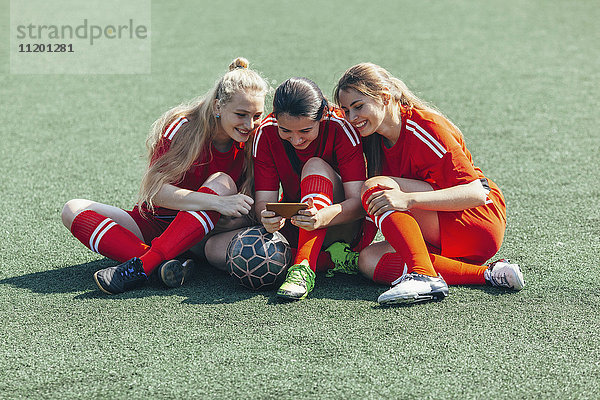 Fröhliche Fußballspieler beim Sitzen auf dem Spielfeld mit Smartphone