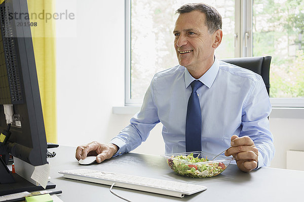 Lächelnder Geschäftsmann bei der Arbeit am Computer und Salat am Schreibtisch im Kreativbüro