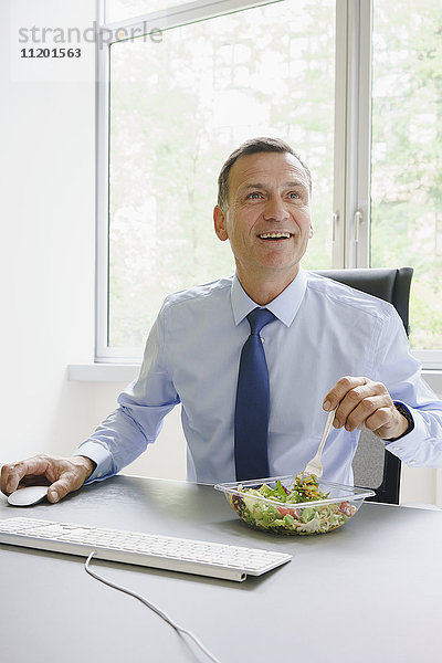 Lächelnder Geschäftsmann mit Salat am Schreibtisch im Kreativbüro