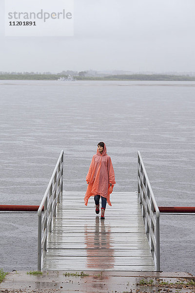 Frau im Regenmantel  die während der Regenzeit auf dem Steg läuft.
