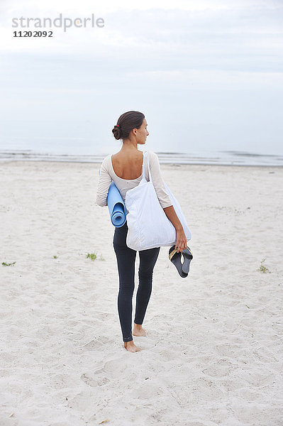 Junge Frau mit Übungsmatte am Strand stehend