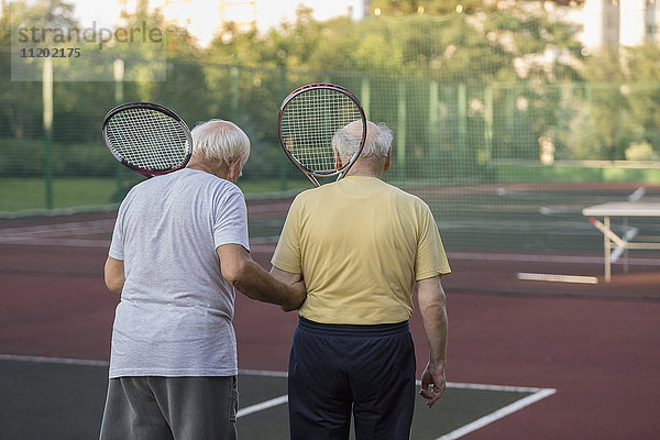 Rückansicht von älteren Freunden  die Tennisschläger tragen  während sie auf dem Spielfeld spazieren gehen.
