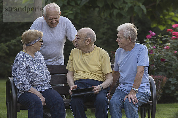 Lächelnde Senior-Freunde beim Gespräch mit dem digitalen Tablett auf der Parkbank