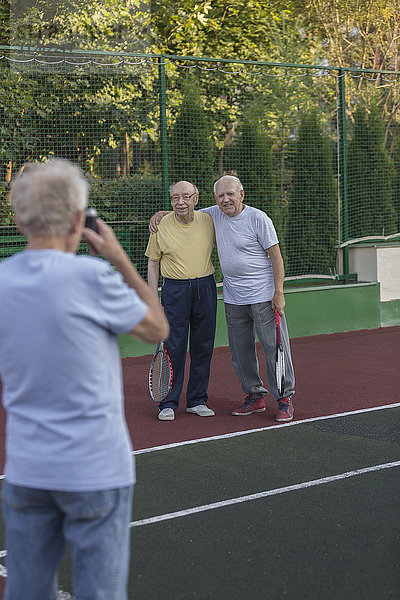 Rückansicht eines Mannes  der Freunde fotografiert  die am Zaun des Tennisplatzes stehen.