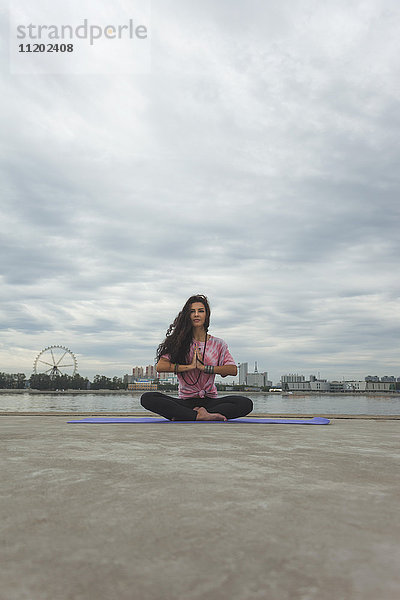 Junge Frau  die Yoga in Gebetsposition gegen den Fluss in der Stadt praktiziert.