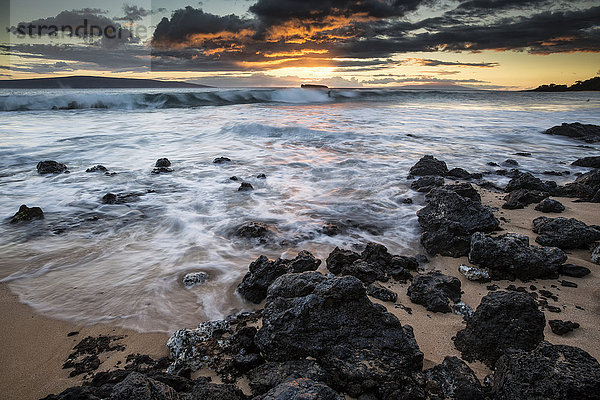 Wasser  das auf die schwarzen Felsen und den Sand am Strand bei Sonnenuntergang gespült wird; Maui  Hawaii  Vereinigte Staaten von Amerika'.