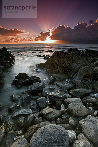 Sonnenstrahlen scheinen über den Wolken  während die Sonne über dem Meer untergeht  mit Felsen entlang der Küste im Vordergrund; Hawaii  Vereinigte Staaten von Amerika'.