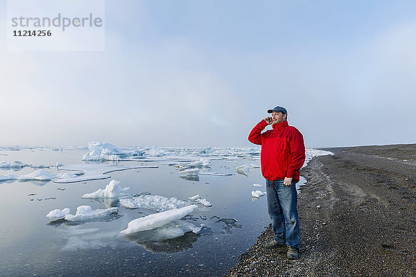 Ein Mann steht an der Küste des Arktischen Ozeans während eines nebligen Sonnenuntergangs und spricht mit seinem Handy  während im Hintergrund Meereis schwimmt; Barrow; North Slope; Arktisches Alaska; USA; Sommer
