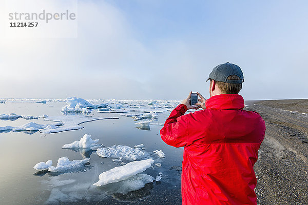 Ein Mann steht an der Küste des Arktischen Ozeans während eines nebligen Sonnenuntergangs und macht ein Foto mit seinem Handy  während das Meereis im Hintergrund schwimmt; Barrow; North Slope; Arktisches Alaska; USA; Sommer