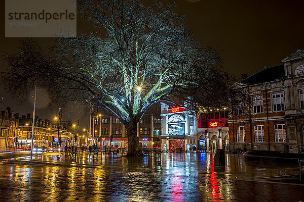 Lichter beleuchten Gebäude und spiegeln sich auf der nassen Straße  Brixton  South London; London  England'.