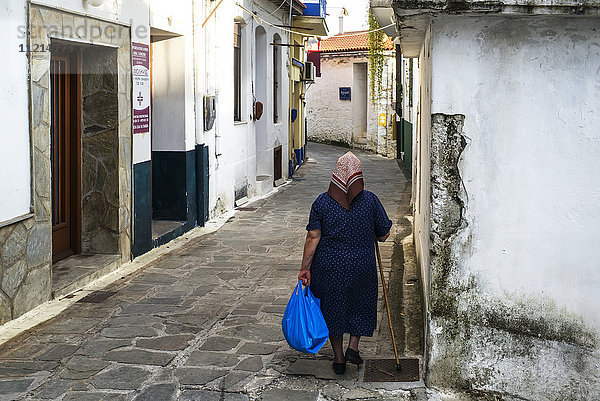 Eine ältere Frau geht mit einem Stock und einer blauen Tasche eine schmale Straße zwischen Häusern auf einer griechischen Insel entlang; Glossa  Skiathos  Griechenland'.