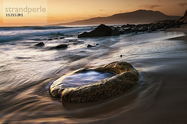 Wasser wird auf den Sand und einen Felsen am Strand gespült  während der Himmel bei Sonnenuntergang orange leuchtet; Tarifa  Cadiz  Andalusien  Spanien'.