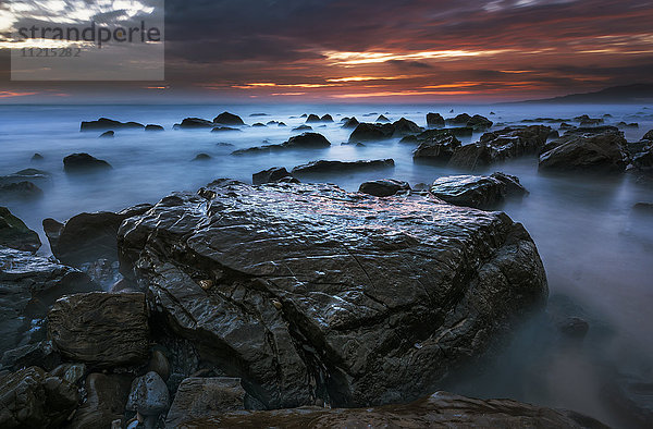 Wasser umgibt die Felsen am Strand  und die Wolken leuchten bei Sonnenuntergang orange und rosa über dem Horizont; Tarifa  Cádiz  Andalusien  Spanien'.