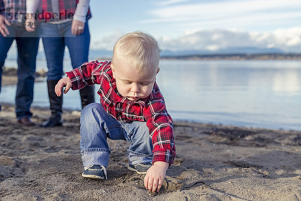 Ein Kleinkind macht sich beim Spielen mit Sand am Strand die Hände schmutzig  während seine Eltern im Hintergrund zusehen; Surrey  British Columbia  Kanada'.