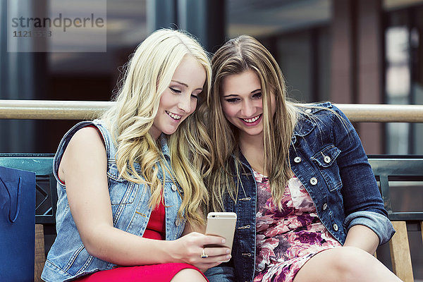 Zwei hübsche junge Frauen beim Einkaufen  die eine Pause einlegen  um sich auf einem Smartphone über soziale Medien zu informieren; Edmonton  Alberta  Kanada'.