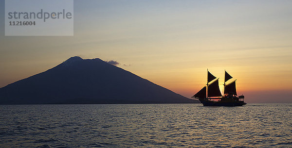 Blick auf das Meer mit einer Yacht mit Segeln und einem Vulkan  der sich gegen die untergehende Sonne abhebt