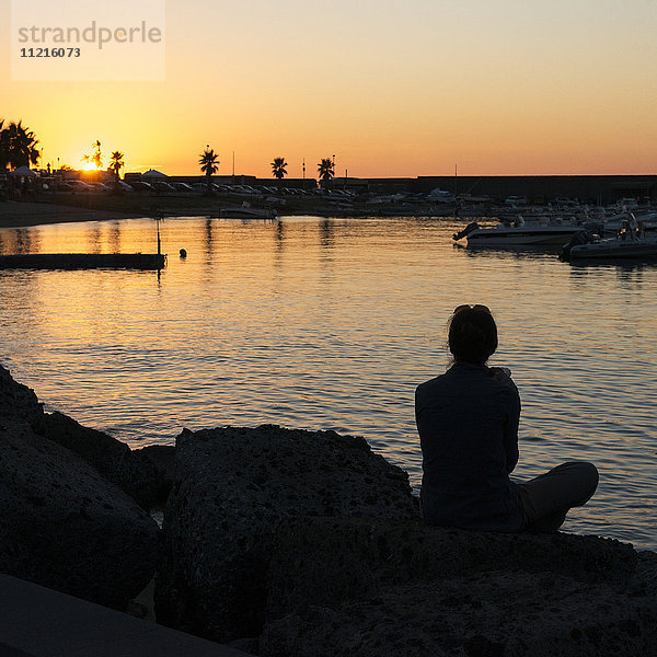 Silhouette einer Frau  die am Ufer sitzt und bei Sonnenuntergang auf einen Hafen blickt; Forio  Ischia  Kampanien  Italien'.