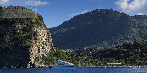 Eine Yacht neben einer hohen Klippe und einer bergigen Landschaft auf der Insel Ischia; Ischia  Kampanien  Italien