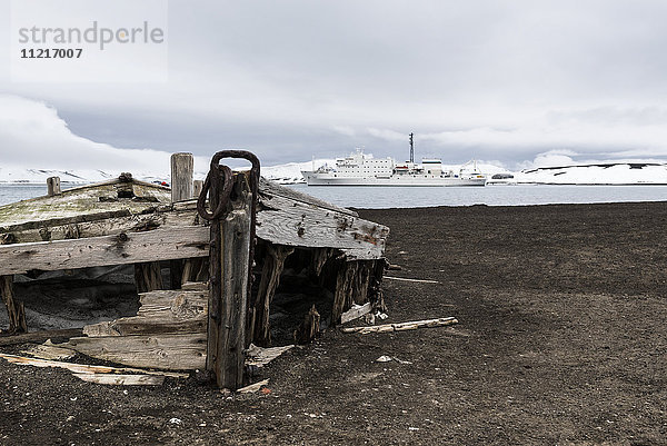 Altes Holzboot verlassen am Ufer einer Walfangstation mit einem Kreuzfahrtschiff im Wasser; Deception Island  South Shetlands  Antarktis