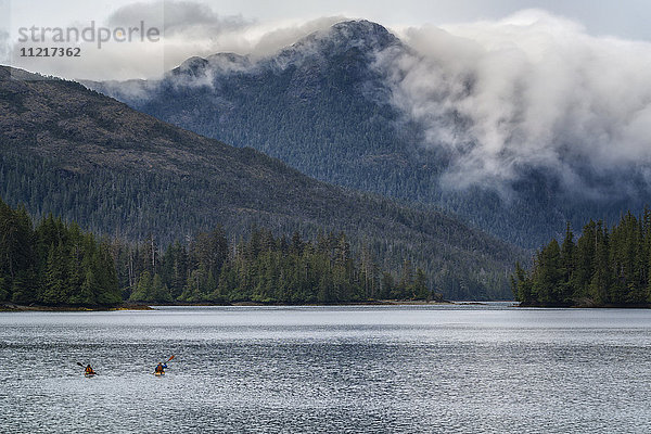 Zwei Kajakfahrer paddeln in Richtung Festland  während Wolken über die Küstenberge ziehen; Haida Gwaii  British Columbia  Kanada'.